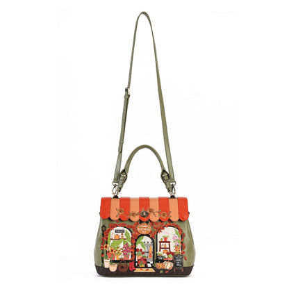 A structured olive green handbag with a colorful, whimsical storefront illustration on the front, featuring a terracotta awning, floral details, and a long shoulder strap, set against a plain white background.
