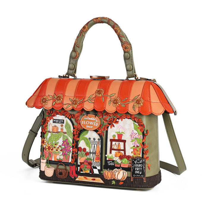 A structured handbag with a house-shaped autumn flower shop design, featuring a striped orange and peach awning with embroidered sunflowers, olive green body, and detailed storefront illustrations including pumpkins, flowers, and a chalkboard sign.