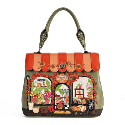An olive green handbag with a scalloped orange and peach flap, decorated with autumn-themed embroidery including pumpkins, flowers, and storefront windows. The bag has dual top handles and a central metal clasp, photographed against a solid white background.