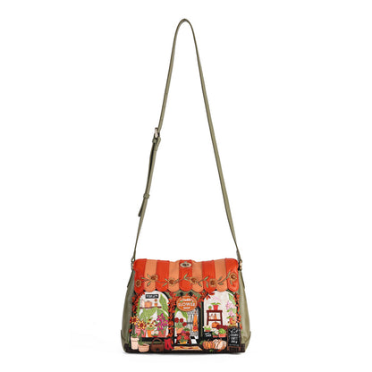 A shoulder bag with a wide, colorful patchwork front panel depicting vintage market scenes and a striped orange-red top flap, paired with a slim olive green strap, shown against a plain white background.