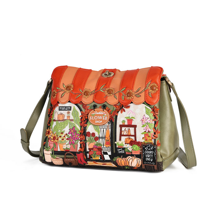 A shoulder bag with a vibrant autumn-themed illustration of a flower shop window on the front, featuring orange and peach scalloped leather trim with embroidered flowers, olive green side panels, and a matching adjustable shoulder strap, set against a plain white background.