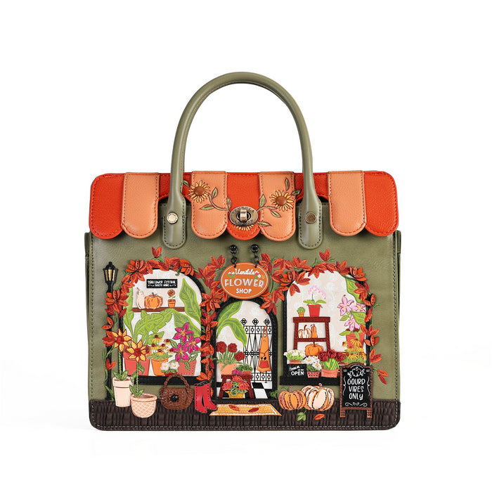 An olive green handbag with a highly detailed, colorful autumn-themed illustration on the front depicting flower shop windows, pumpkins, and fall foliage, complete with orange and peach flap details and top handles.