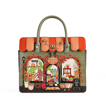 An olive green handbag with a highly detailed, colorful autumn-themed illustration on the front depicting flower shop windows, pumpkins, and fall foliage, complete with orange and peach flap details and top handles.