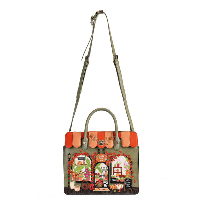 An olive green tote bag with a long shoulder strap, featuring a bright orange scalloped trim and a detailed colorful illustration of a cozy autumn storefront on the front.