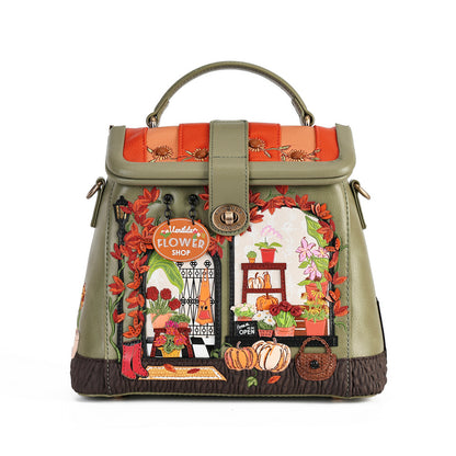 An olive green handbag with a top handle, featuring a detailed embroidered design of a flower shop storefront with pumpkins, flowers, and autumn leaves.