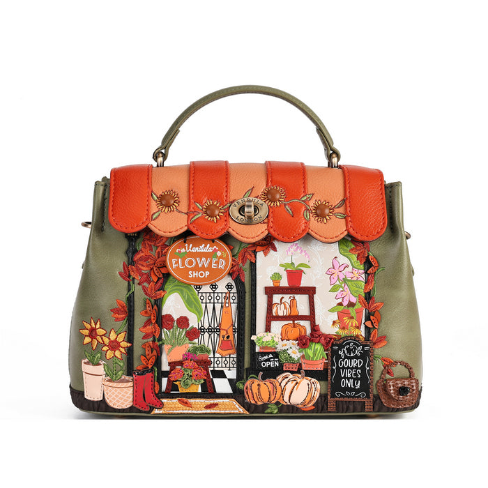 A structured olive green handbag with a top handle, featuring a prominent orange flap and a highly detailed, colorful embroidered autumn flower shop scene on the front.
