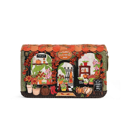 A rectangular zip-around clutch bag featuring a highly detailed, colorful embroidered design of a flower shop storefront with autumn elements like pumpkins, flowers, and a scalloped orange roof, shown against a plain white background.