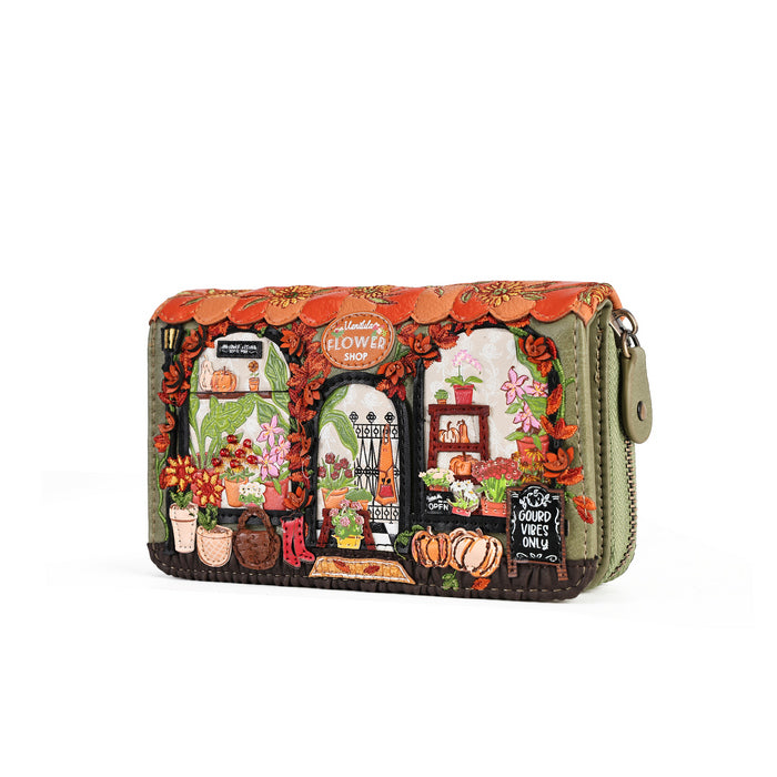 A rectangular zip-around wallet with a detailed autumn-themed illustration on the front, featuring pumpkins, flowers, a flower shop sign, and a green fabric border, shown on a plain white background.