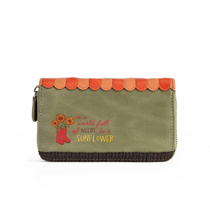 An olive green zip-around wallet with a scalloped orange and peach trim along the top, a dark brown stitched bottom edge, and embroidered text reading 'in a world full of weeds, be a sunflower' with sunflowers and red boots.