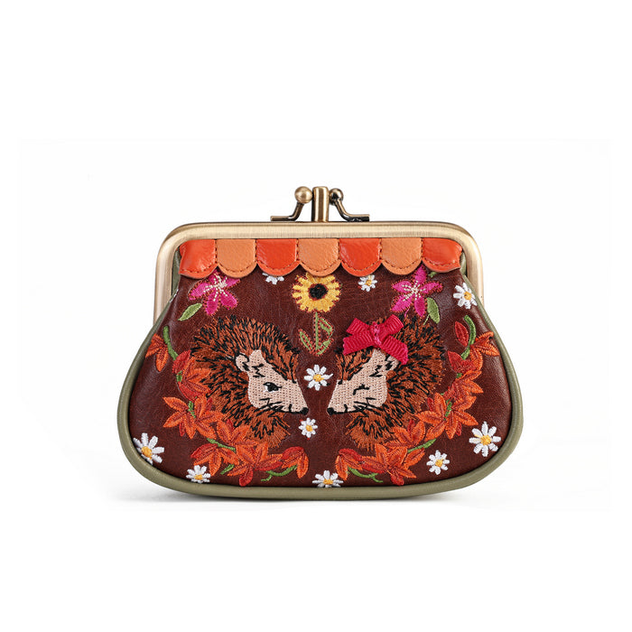 A small kiss-lock clutch purse with a dark brown background and detailed embroidery of two hedgehogs, orange leaves, pink flowers, and white daisies, framed by a scalloped orange border and a gold-tone metal clasp.