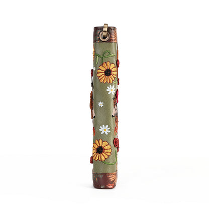 A vertical olive green leather sleeve decorated with embroidered sunflowers, white daisies, red ladybugs, and green leaves, featuring brown leather trim and a brass ring at the top.