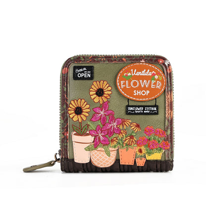 A small zip-around wallet with a floral patchwork design, featuring embroidered flowers in pots, decorative stickers including 'Vendetta FLOWER SHOP', and a dark brown ruffled trim.