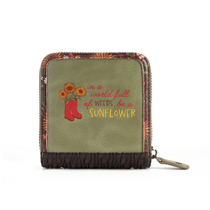 An olive green zip-around wallet with a floral patterned border, a dark brown gathered bottom, and embroidered text reading 'in a world full of weeds, be a SUNFLOWER' alongside red boots and yellow sunflowers.