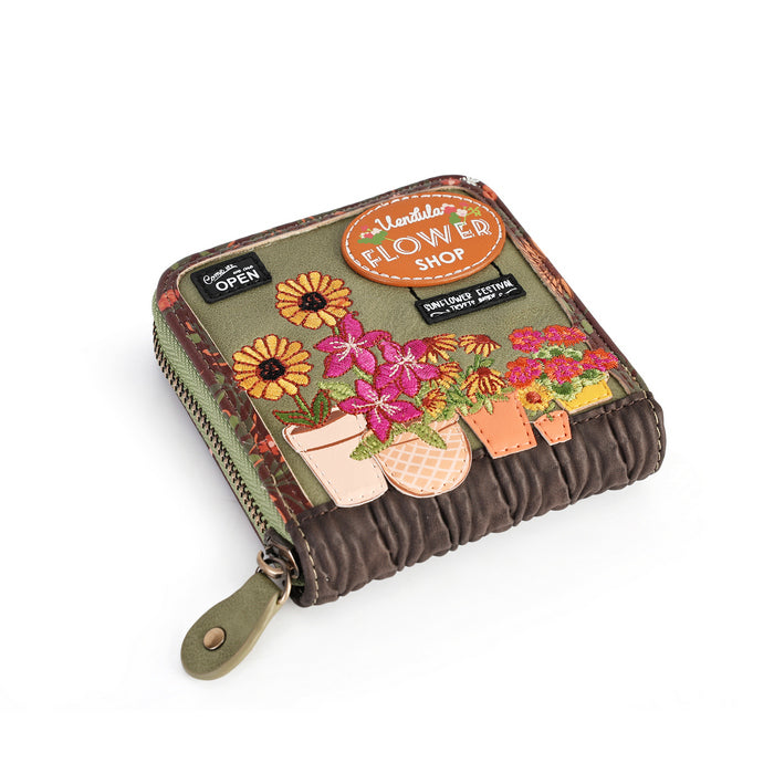 A small zip-around wallet with a quilted brown base and an olive green top panel decorated with embroidered flowers, potted plants, and a circular 'Flower Shop' patch. A black tab on the left reads 'GREATLY OPEN'.