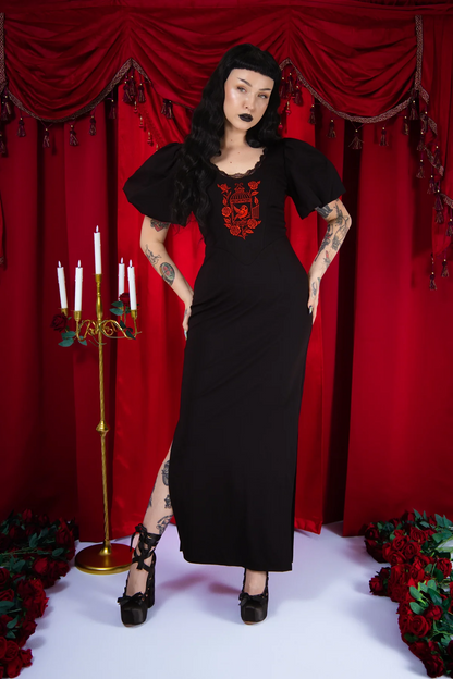 Woman in a black dress with red embroidery standing in front of red curtains with candles and flowers.