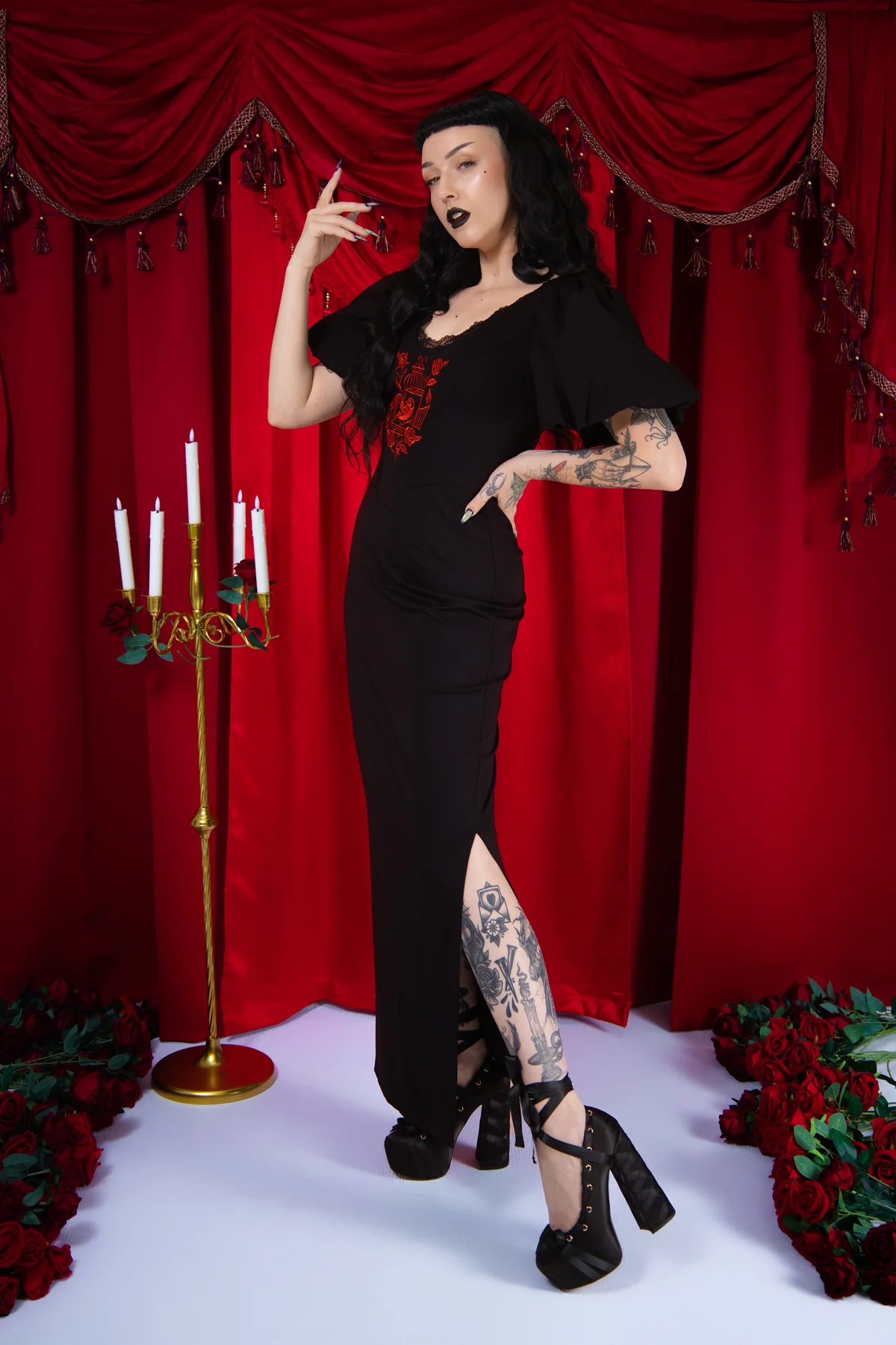 Woman in a black outfit posing in front of red curtains with candles and floral decorations.
