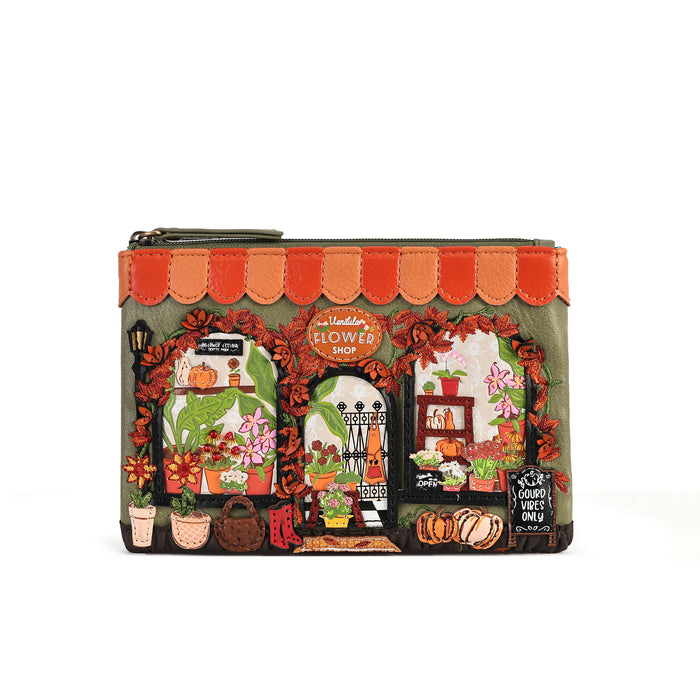 A small zip-around pouch with a printed illustration of a flower shop facade. The pouch has an olive green base, a scalloped orange and peach awning, and detailed storefront windows showing flowers, pumpkins, plants, and a chalkboard sign, all framed by red floral garlands.