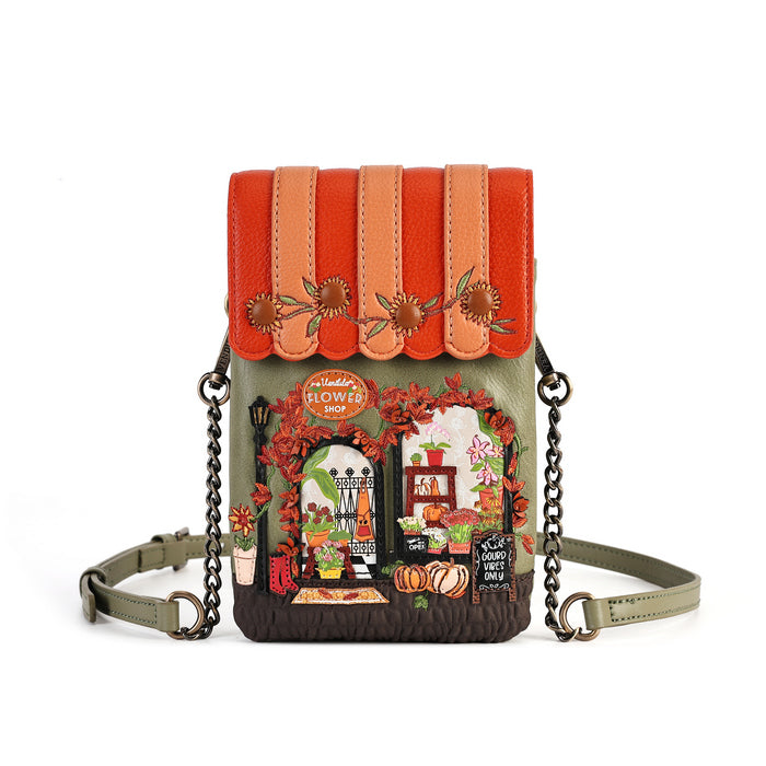 A small crossbody phone bag with a colorful autumn-themed storefront design. The top flap features orange and peach faux-leather panels with embroidered sunflowers, while the main body is olive green with a detailed embroidered shop scene below.