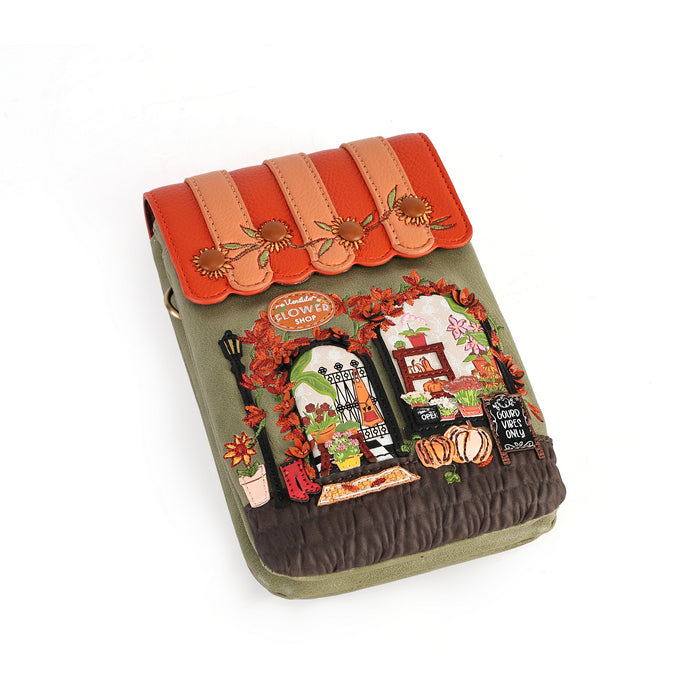 A small quilted fabric crossbody bag with a whimsical embroidered flower shop design on the front flap, featuring a peach and rust faux-leather top flap with embroidered flowers, an olive green body, and a dark brown quilted base.