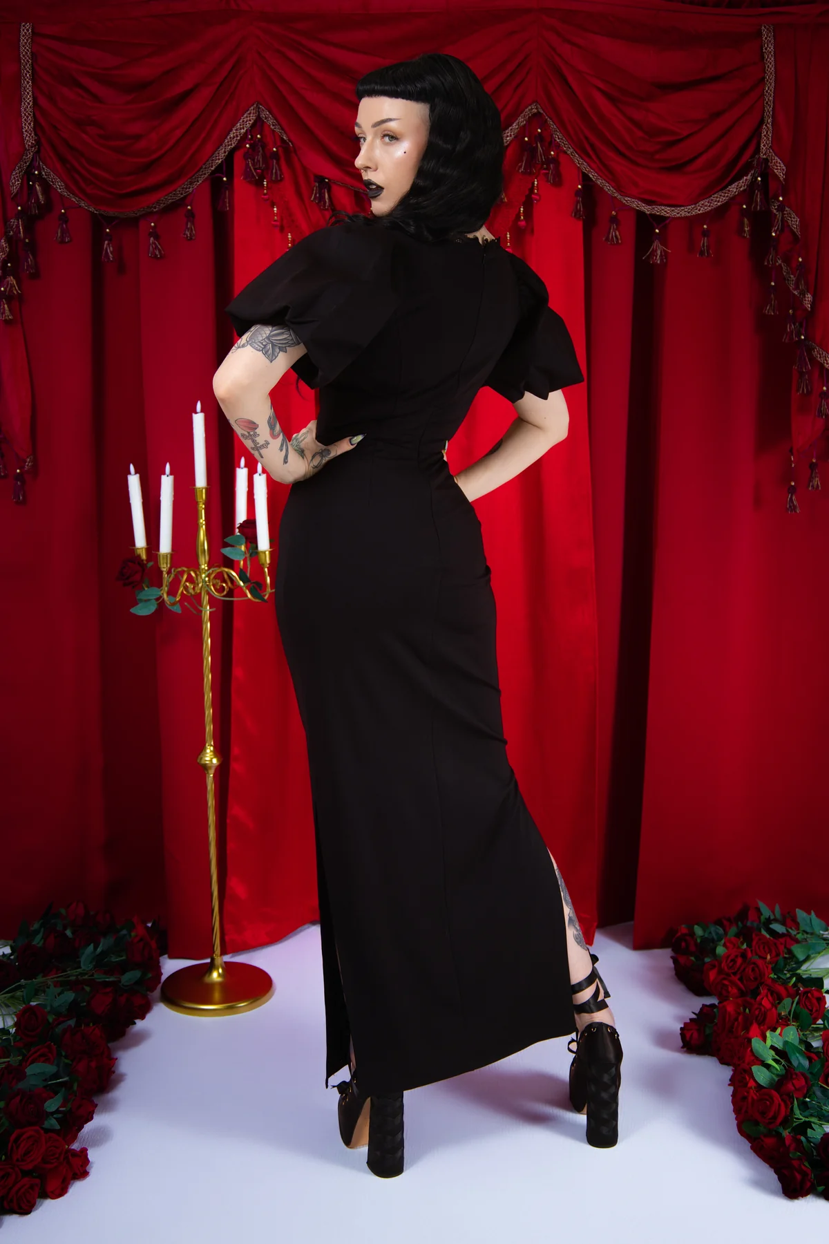 Woman in a black dress posing in front of red curtains with candles and flowers.