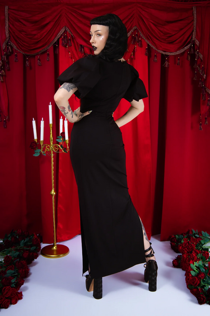 Woman in a black dress posing in front of red curtains with candles and flowers.