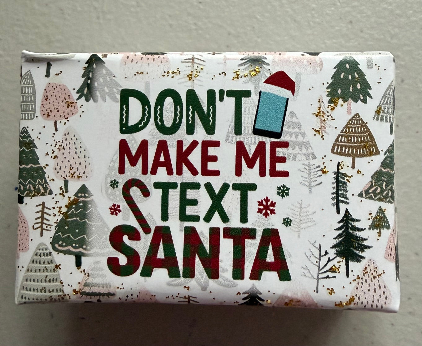 A square gift box wrapped in white holiday-themed paper featuring the text 'DON'T MAKE ME TEXT SANTA' in dark green and red lettering, surrounded by illustrated Christmas trees and snowflakes.