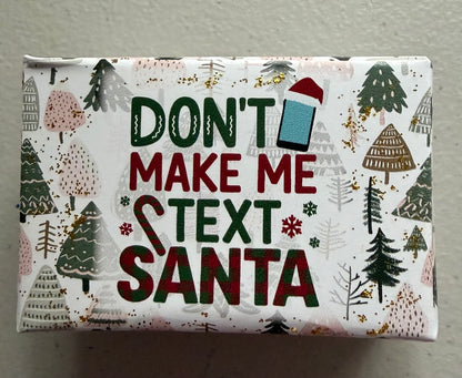 A square gift box wrapped in white holiday-themed paper featuring the text 'DON'T MAKE ME TEXT SANTA' in dark green and red lettering, surrounded by illustrated Christmas trees and snowflakes.