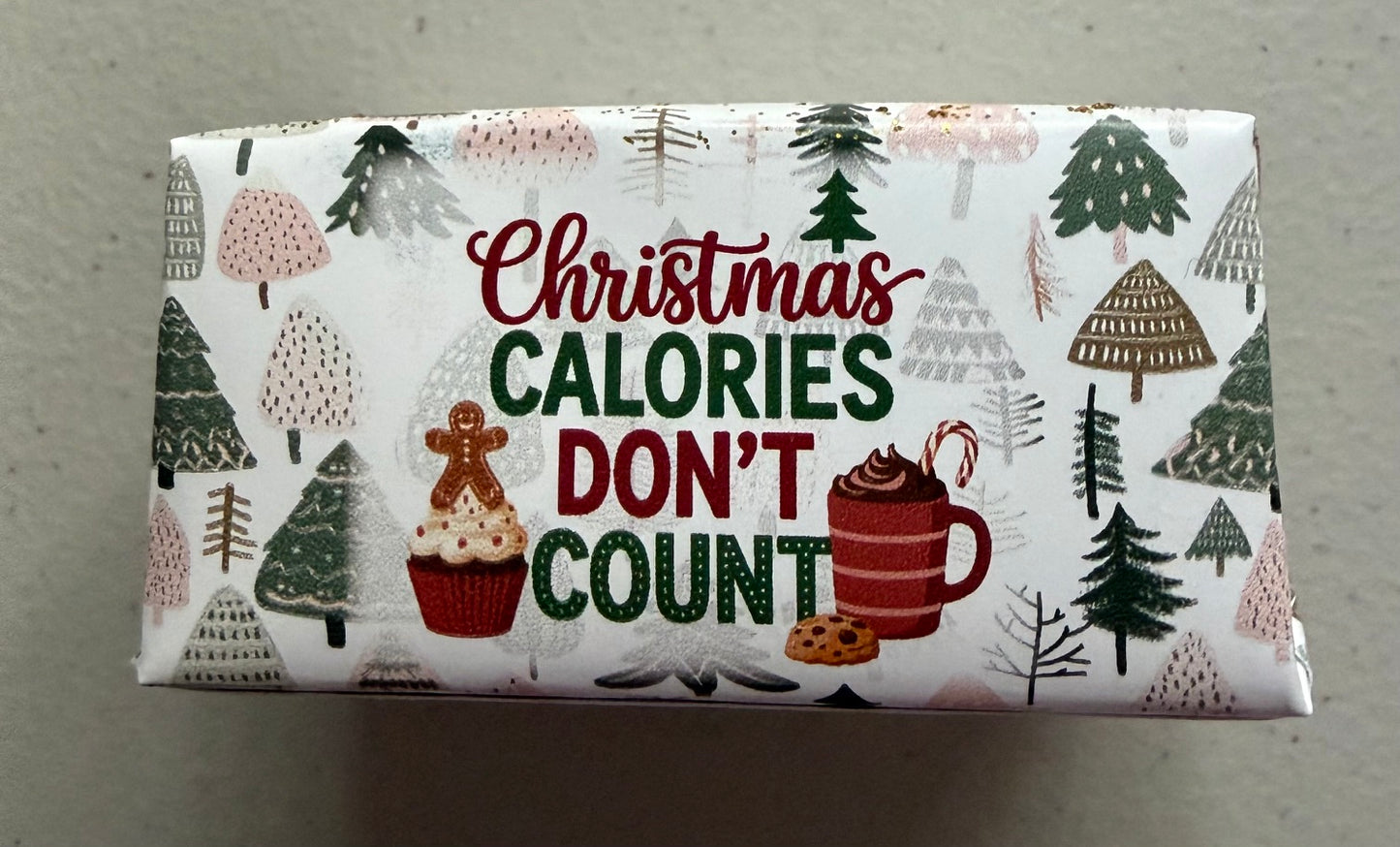 A wrapped candy bar featuring a festive Christmas design with the text 'Christmas CALORIES DON'T COUNT' in red and green lettering, surrounded by illustrations of pine trees, a gingerbread man, a cupcake, and a mug of hot chocolate on a white background.