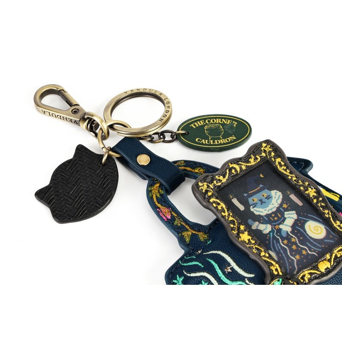 A decorative keychain set featuring a black textured cat-shaped charm, a gold-toned lobster clasp and split ring, a green oval tag reading 'THE CORNE'S CAULDRON', and a navy blue fabric charm with gold trim and a blue cauldron illustration.