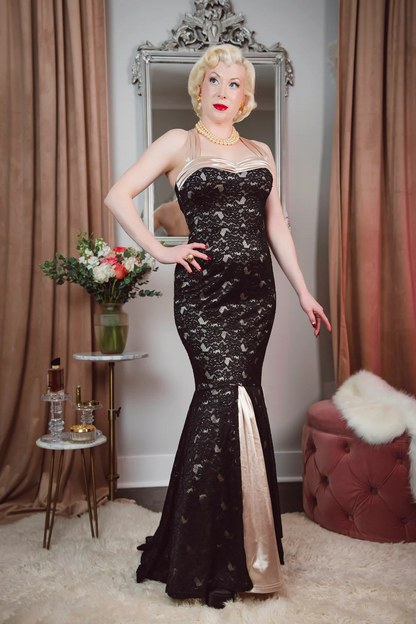 Woman in a black lace evening gown standing in a room with curtains and a mirror.