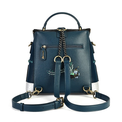 A dark teal blue leather backpack with gold-tone hardware, featuring a top handle, a detachable shoulder strap, a front zip pocket, and a small embroidered cartoon character on the lower right side.