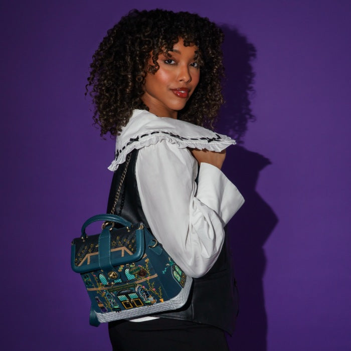 A woman with curly dark hair models a teal and black embroidered handbag with gold details, styled with a white ruffled shirt against a solid purple background.