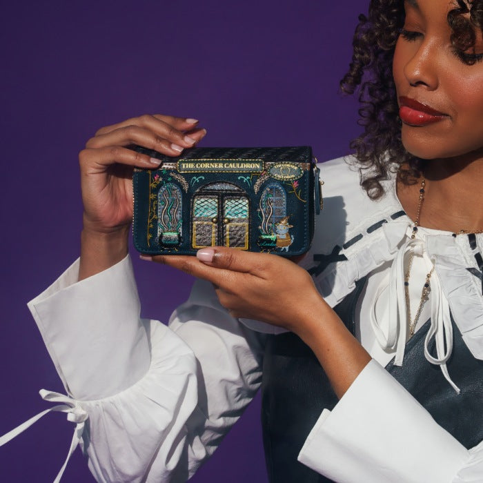 A woman in a white blouse holds up a small decorative coin purse against a solid purple background. The purse is dark teal with intricate gold, green, and blue embroidery depicting a cathedral facade, and has a gold plaque at the top reading 'THE CATHEDRAL CALENDAR'.
