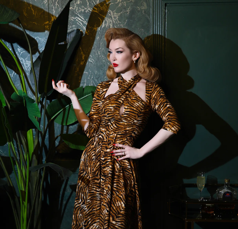 Woman in a tiger-striped dress holding a large leaf in a dimly lit room with plants and a bottle.