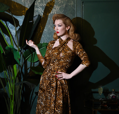 Woman in a tiger-striped dress holding a large leaf in a dimly lit room with plants and a bottle.