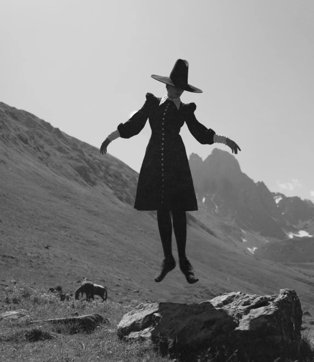 Person in a coat and hat floating above a mountain landscape with a cow and sheep.