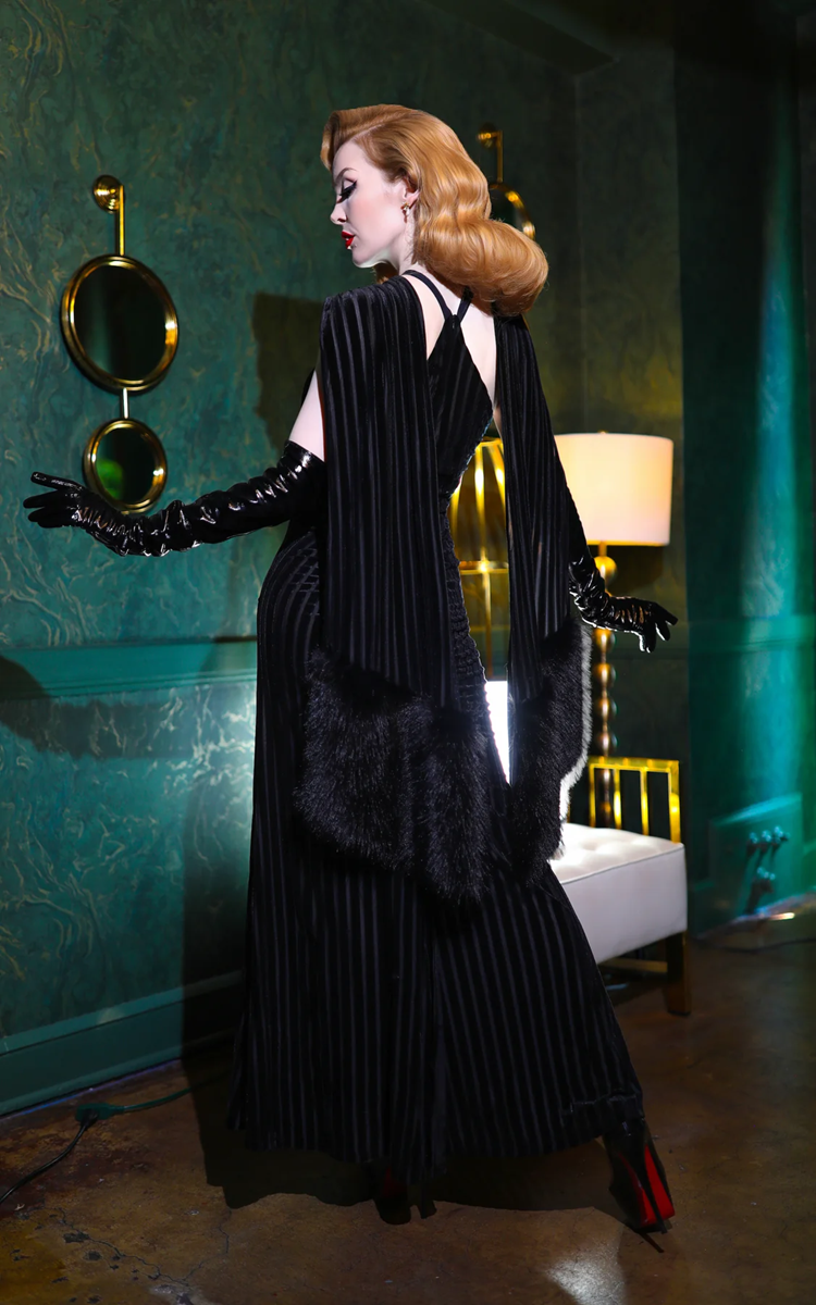 Woman in a black evening gown with gloves holding a mirror in a dimly lit room.