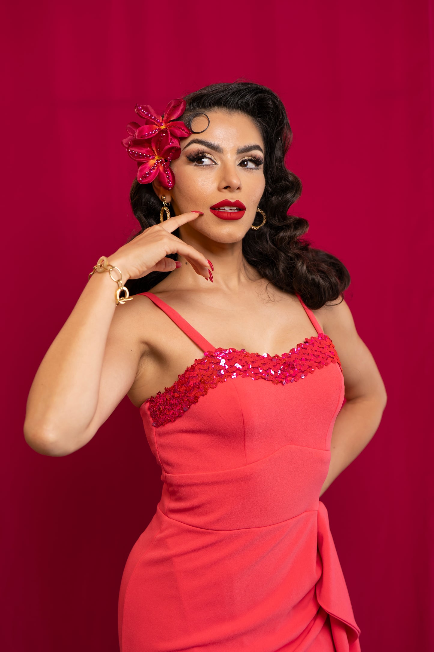A woman posing in a retro-style coral pink dress with sequined detailing on the bust, set against a solid dark pink background.