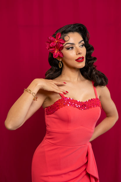 A woman posing in a bright coral pink sequined dress against a solid dark pink background, featuring a vintage-inspired hairstyle with a pink flower accessory.