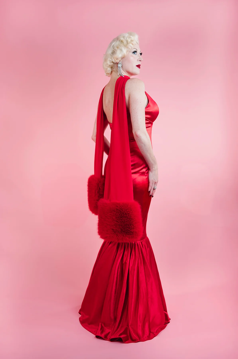 Woman in a red evening gown with fur accents against a pink background