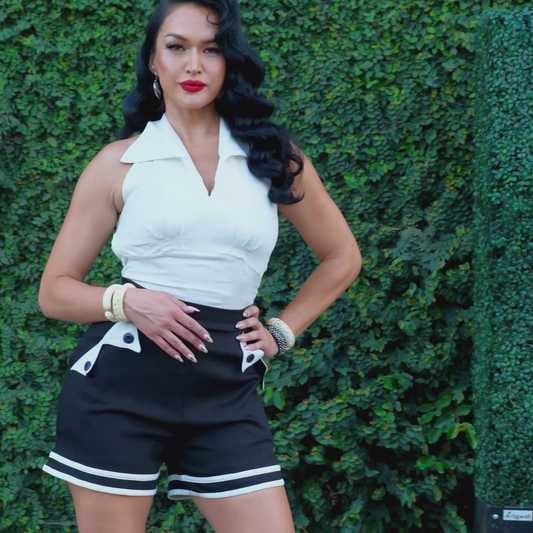 A woman posing in front of a green ivy wall, wearing a white sleeveless collared top and black shorts with white accents.