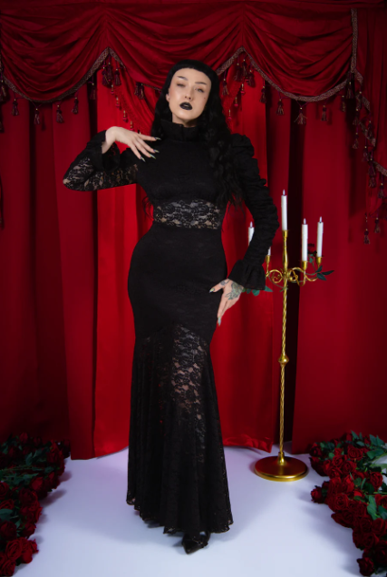 Woman in a black lace dress posing in front of red curtains with a candelabra and flowers.