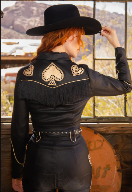 Person wearing a black cowboy hat and black jacket with decorative elements, standing indoors.