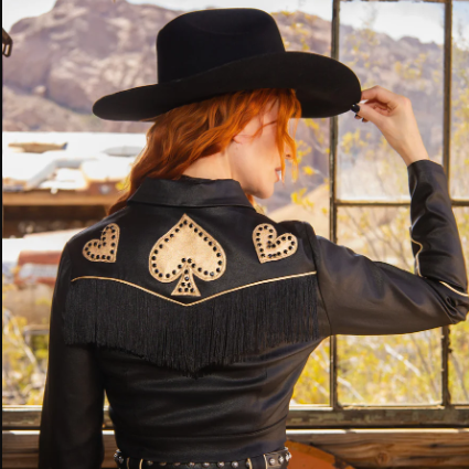 Person wearing a black cowboy hat and a black jacket with heart and spade designs, standing in front of a window with a mountain view.