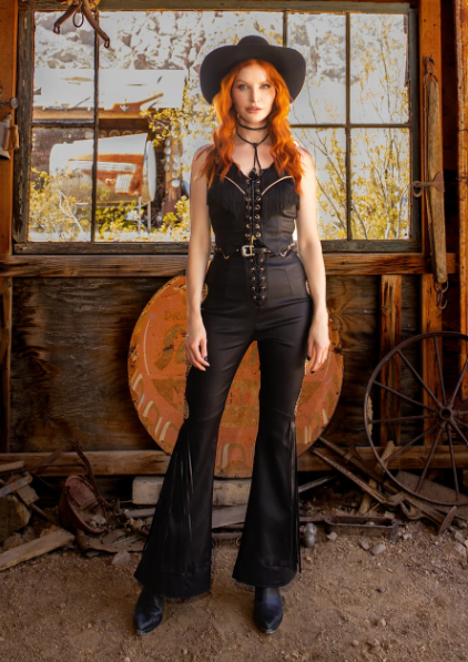 Woman in black leather outfit with red hair and cowboy hat standing in rustic setting