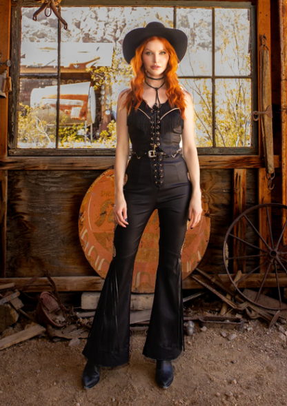 Woman in black leather outfit with red hair and cowboy hat standing in rustic setting