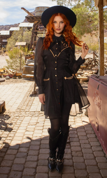 Woman in a black outfit with gold accents standing in a rustic, sunlit outdoor setting.