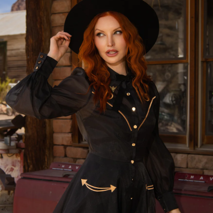 A woman with long red hair wearing a black wide-brim hat, a black long-sleeve dress with gold buttons and trim, and posing in front of a rustic wooden building.