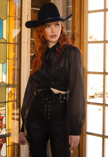 Person wearing a black outfit with a wide-brimmed hat in front of a stained glass window.