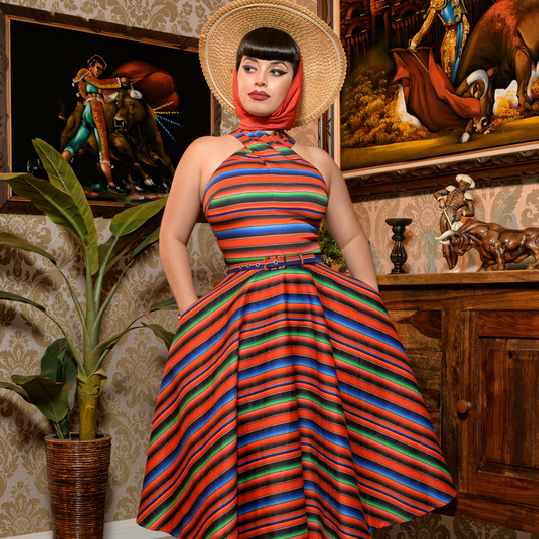 A woman stands indoors wearing a colorful striped halter dress with a flared skirt, a wide-brim straw hat, and a red headscarf. The room has patterned wallpaper, framed artwork, a potted plant, and wooden furniture.